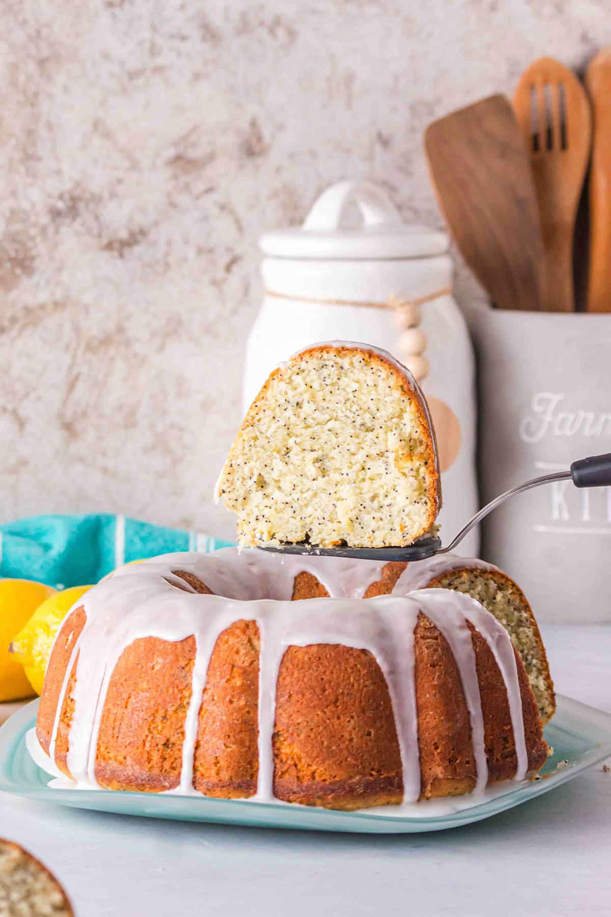 A Lemon Poppy Seed Bundt Cake with a glossy glaze is being served; one slice is lifted with a spatula, revealing the cake&rsquo;s moist, tender interior. Kitchen utensils and a canister sit in the background.