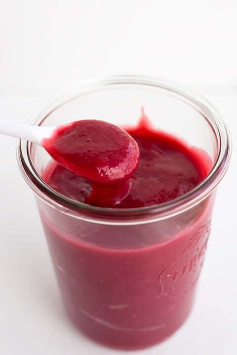 A jar filled with smooth, dark red homemade fruit curd, with a white spoon holding a spoonful above the jar.