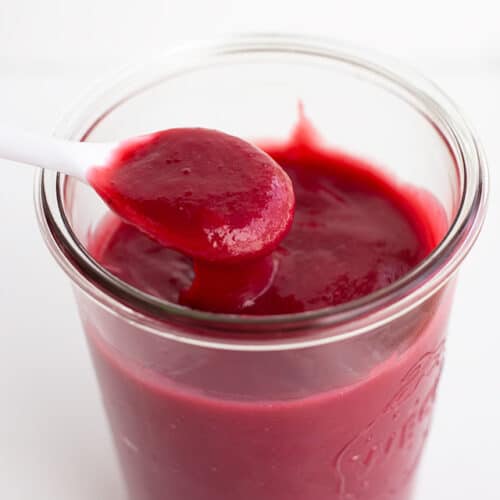 A jar filled with smooth, dark red homemade fruit curd, with a white spoon holding a spoonful above the jar.