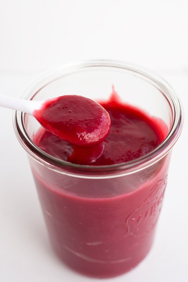A jar filled with smooth, dark red homemade fruit curd, with a white spoon holding a spoonful above the jar.
