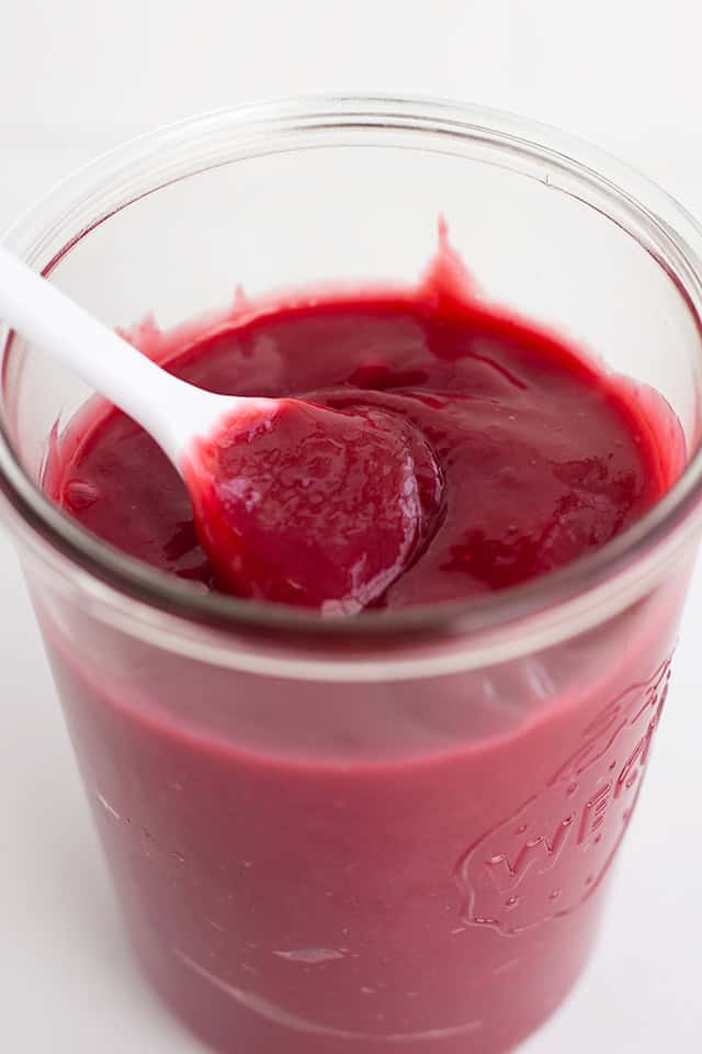 A glass jar filled with thick, deep red Mixed Berry Curd, with a white spoon partially submerged in the luscious mixture.