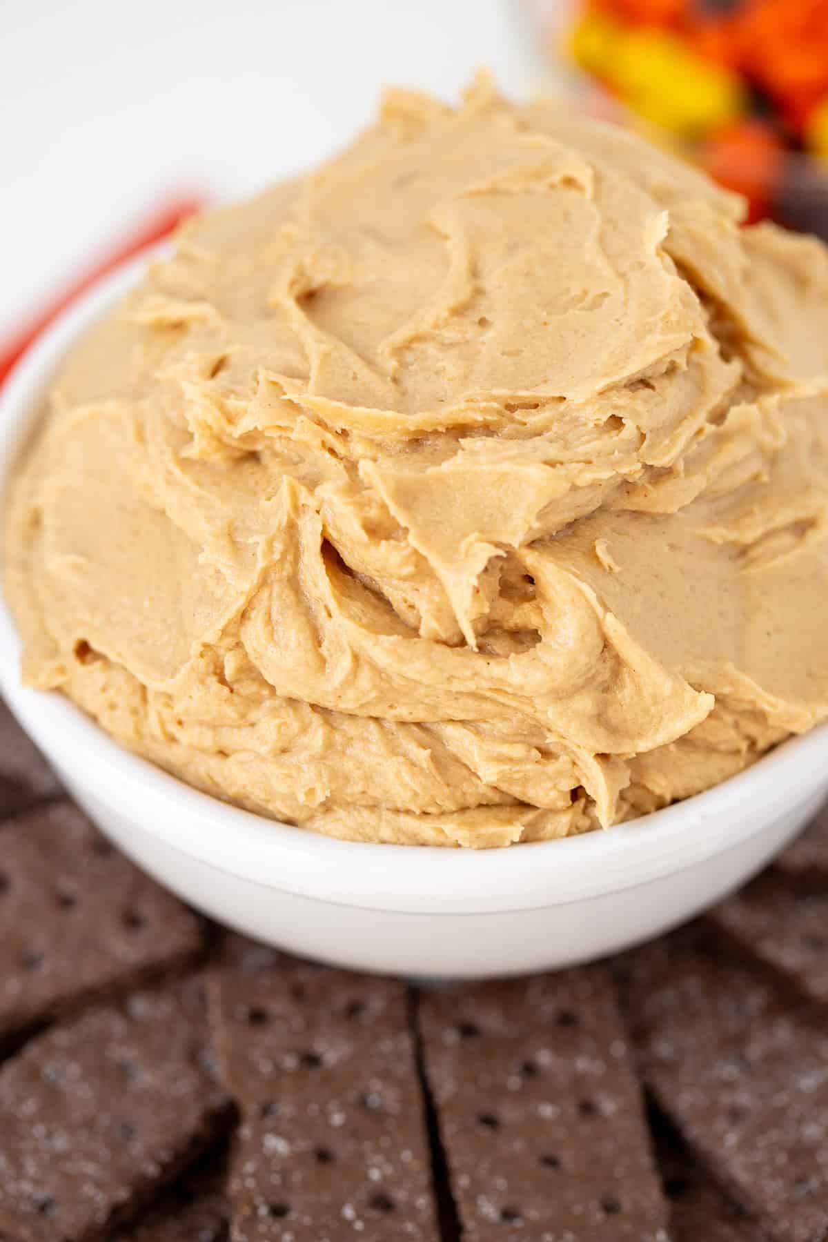 A white bowl filled with creamy Peanut Butter Dip sits next to chocolate cookies, with a blurred bowl of colorful candy in the background.