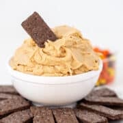 A white bowl filled with creamy Peanut Butter Dip, topped with a single chocolate cracker, surrounded by more chocolate crackers on a white plate.