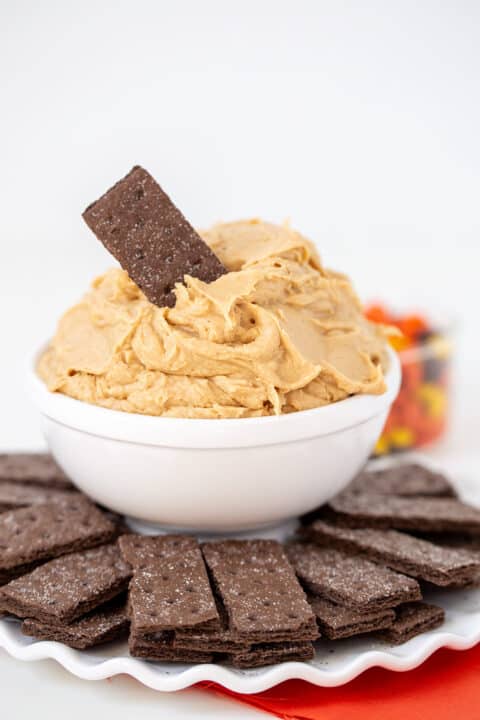 A white bowl filled with creamy Peanut Butter Dip, topped with a single chocolate cracker, surrounded by more chocolate crackers on a white plate.