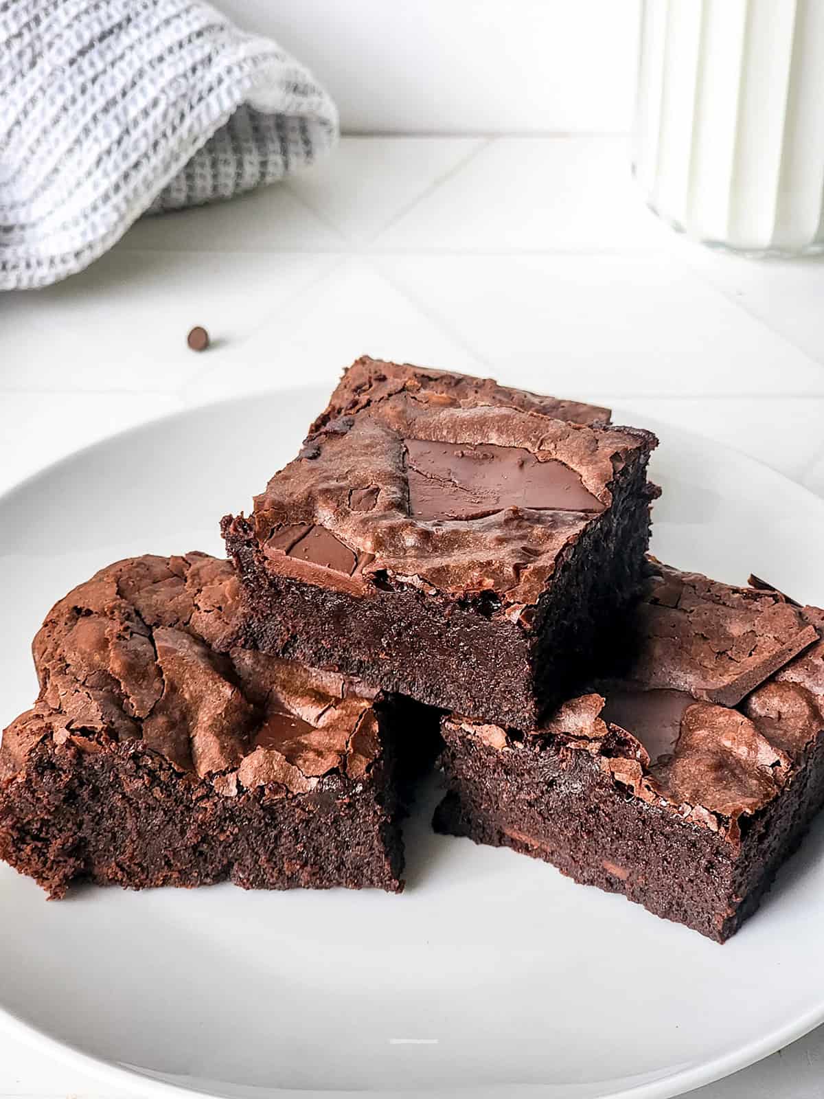 Three rich condensed milk brownies are stacked on a white plate, with a glass of milk and a towel in the background.