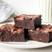Three thick, fudgy sweetened condensed milk brownies are stacked on a white plate, with a glass of milk and a gray kitchen towel in the background.
