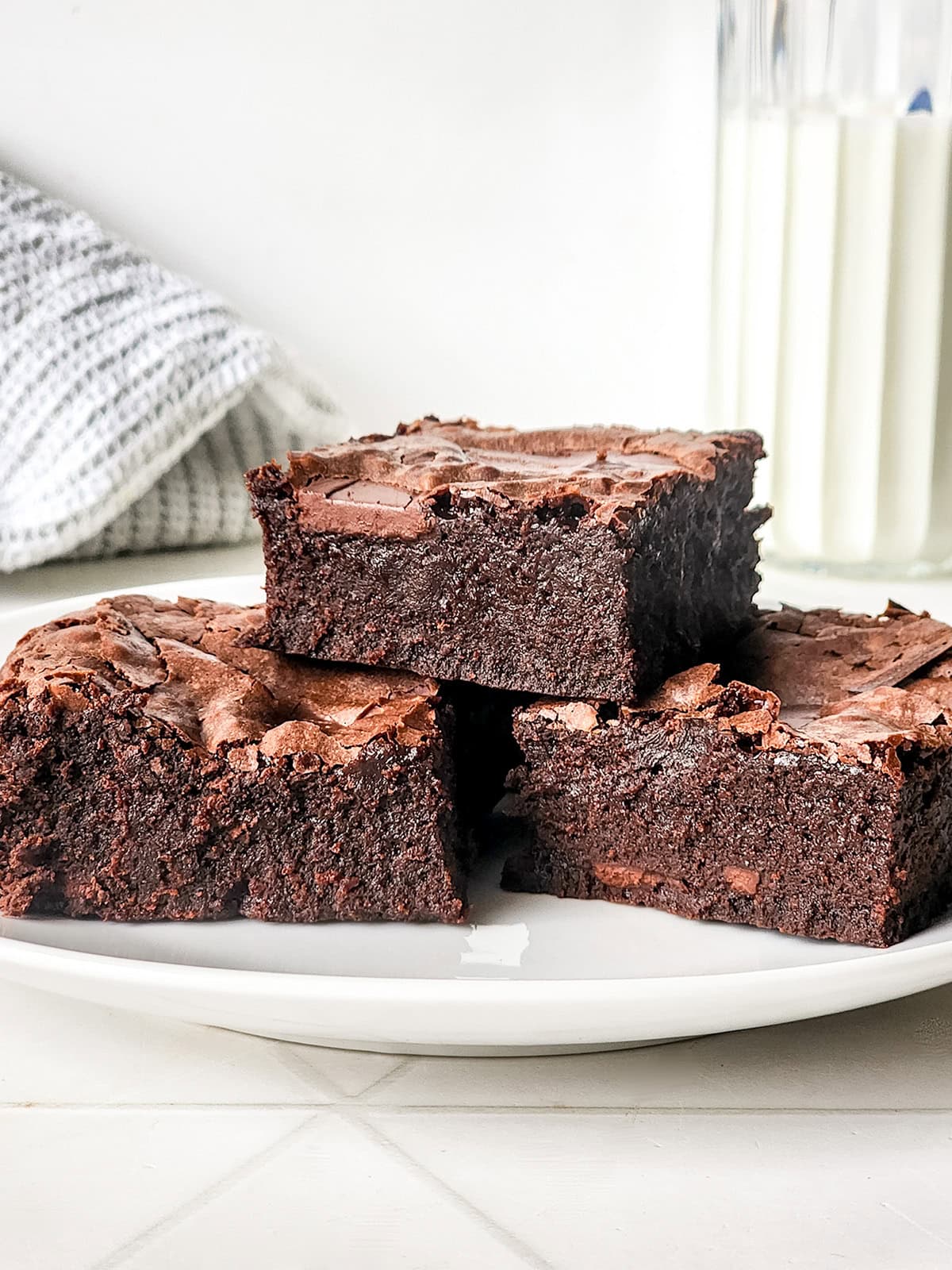Three thick, fudgy sweetened condensed milk brownies are stacked on a white plate, with a glass of milk and a gray kitchen towel in the background.
