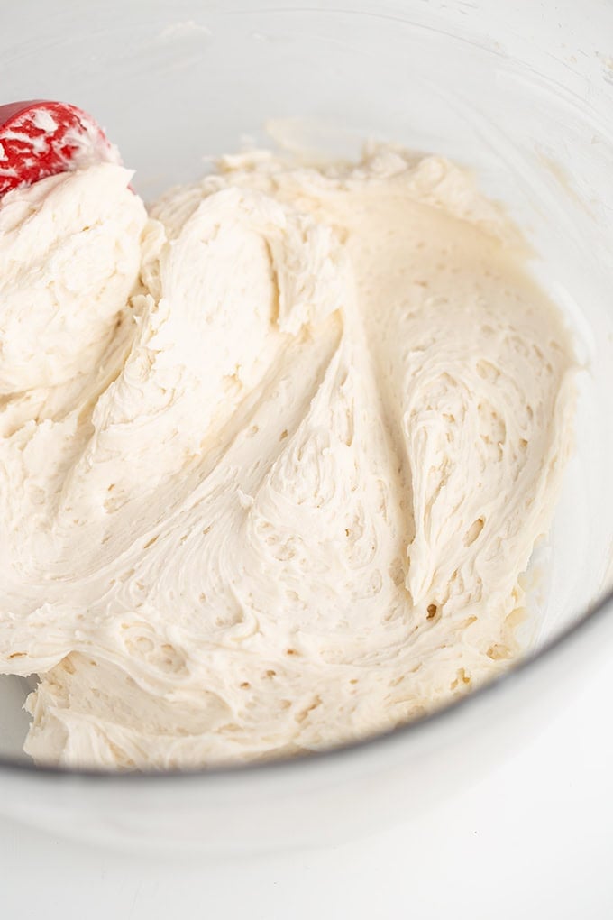 A glass mixing bowl containing creamy, whipped vanilla frosting batter being blended with a red spatula.