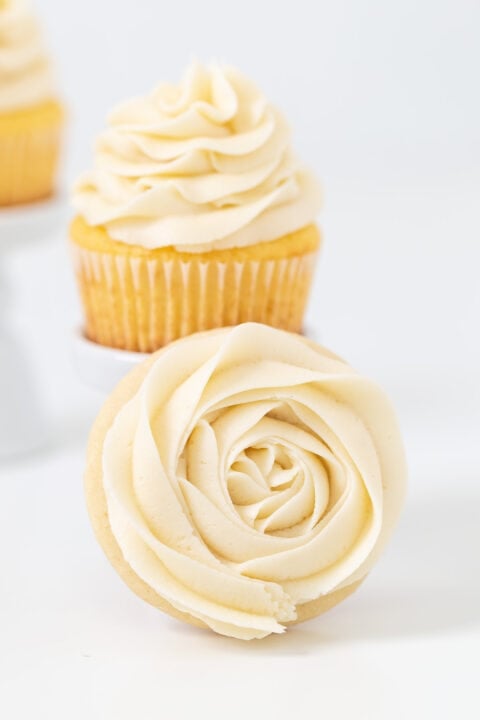 A cupcake topped with swirled vanilla frosting stands behind a cookie adorned with rose-shaped vanilla frosting, both arranged on a white surface.