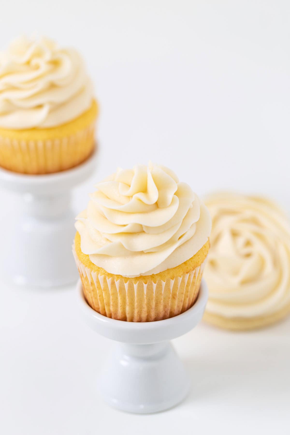 Vanilla cupcakes with swirled homemade frosting on white ceramic stands, with an additional frosted cupcake in the background.