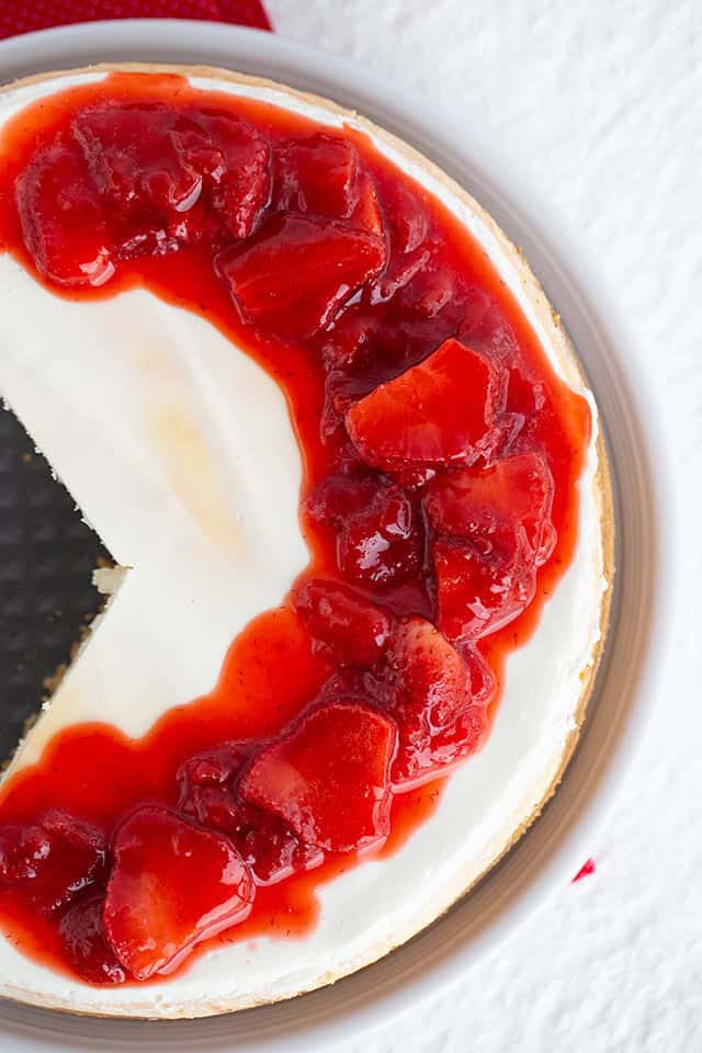 Top view of a Strawberry Cheesecake with a missing slice, topped with strawberry sauce and pieces of strawberries on a white surface.