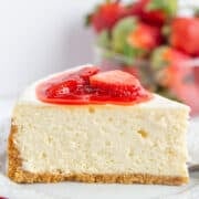A slice of classic Strawberry Cheesecake with a graham cracker crust, topped with strawberry slices and sauce, on a white plate with fresh strawberries in the background—a delightful strawberry dessert.