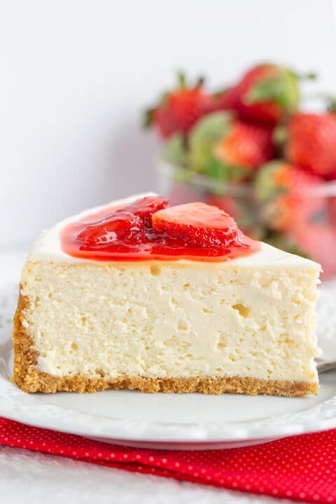 A slice of classic Strawberry Cheesecake with a graham cracker crust, topped with strawberry slices and sauce, on a white plate with fresh strawberries in the background—a delightful strawberry dessert.
