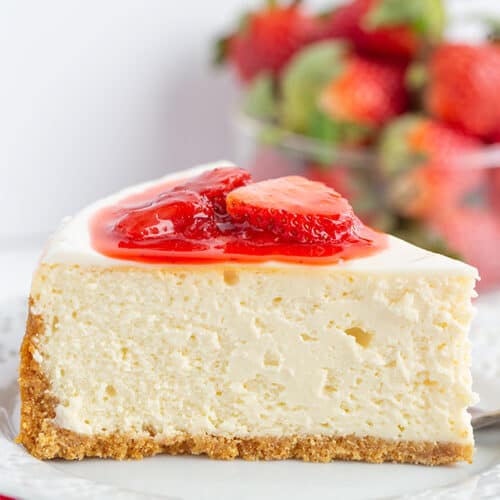 A slice of classic Strawberry Cheesecake with a graham cracker crust, topped with strawberry slices and sauce, on a white plate with fresh strawberries in the background—a delightful strawberry dessert.