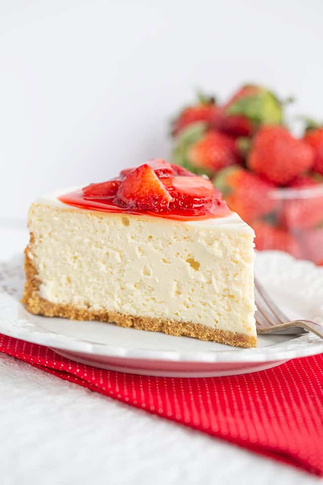A slice of strawberry cheesecake topped with sweet strawberry sauce sits on a white plate, with fresh strawberries in the background—a perfect strawberry dessert.