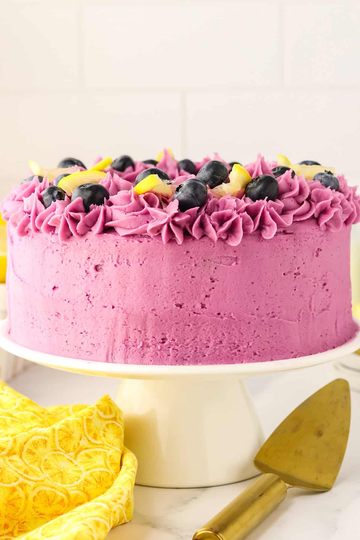 A round Blueberry Lemon Cake with bright purple frosting, decorated with blueberries and lemon slices on top, sits on a white cake stand next to a cake server and a yellow patterned cloth.