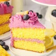 A slice of Blueberry Lemon Cake with purple frosting, topped with a blueberry and a lemon wedge, sits on a plate surrounded by fresh blueberries and lemon slices.