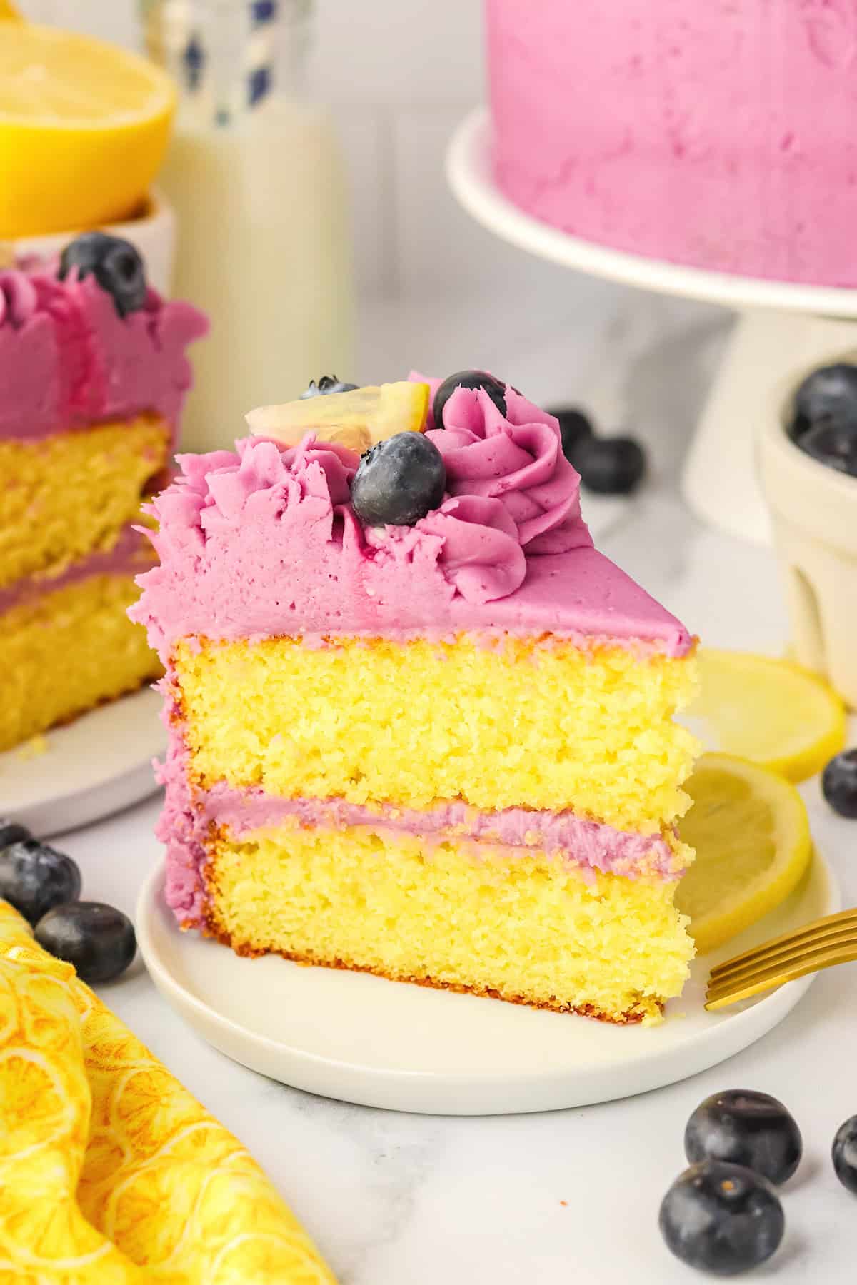 A slice of Blueberry Lemon Cake with purple frosting, topped with a blueberry and a lemon wedge, sits on a plate surrounded by fresh blueberries and lemon slices.