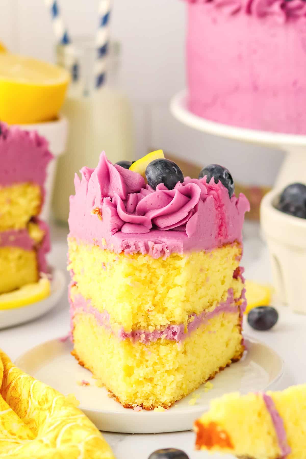 A slice of blueberry lemon cake with purple frosting, topped with blueberries and a lemon slice, sits on a plate in front of the remaining cake.
