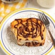 A chocolate chip cinnamon roll with chocolate drizzle sits on a white plate, next to a fork, a glass of milk, and a bowl of chocolate chips atop a yellow checkered cloth.