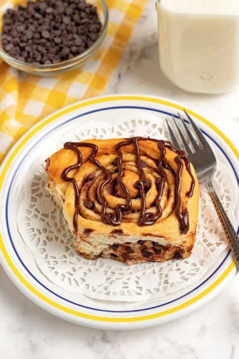 A chocolate chip cinnamon roll with chocolate drizzle sits on a white plate, next to a fork, a glass of milk, and a bowl of chocolate chips atop a yellow checkered cloth.