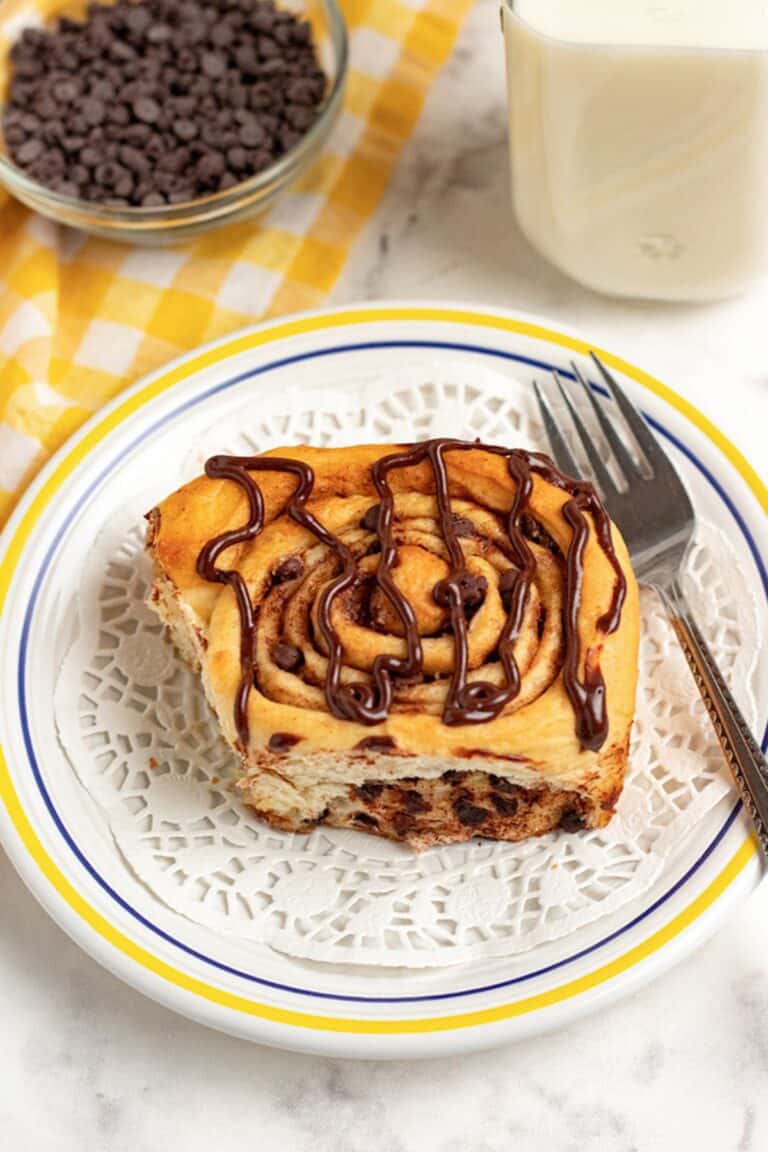 A chocolate chip cinnamon roll with chocolate drizzle sits on a white plate, next to a fork, a glass of milk, and a bowl of chocolate chips atop a yellow checkered cloth.