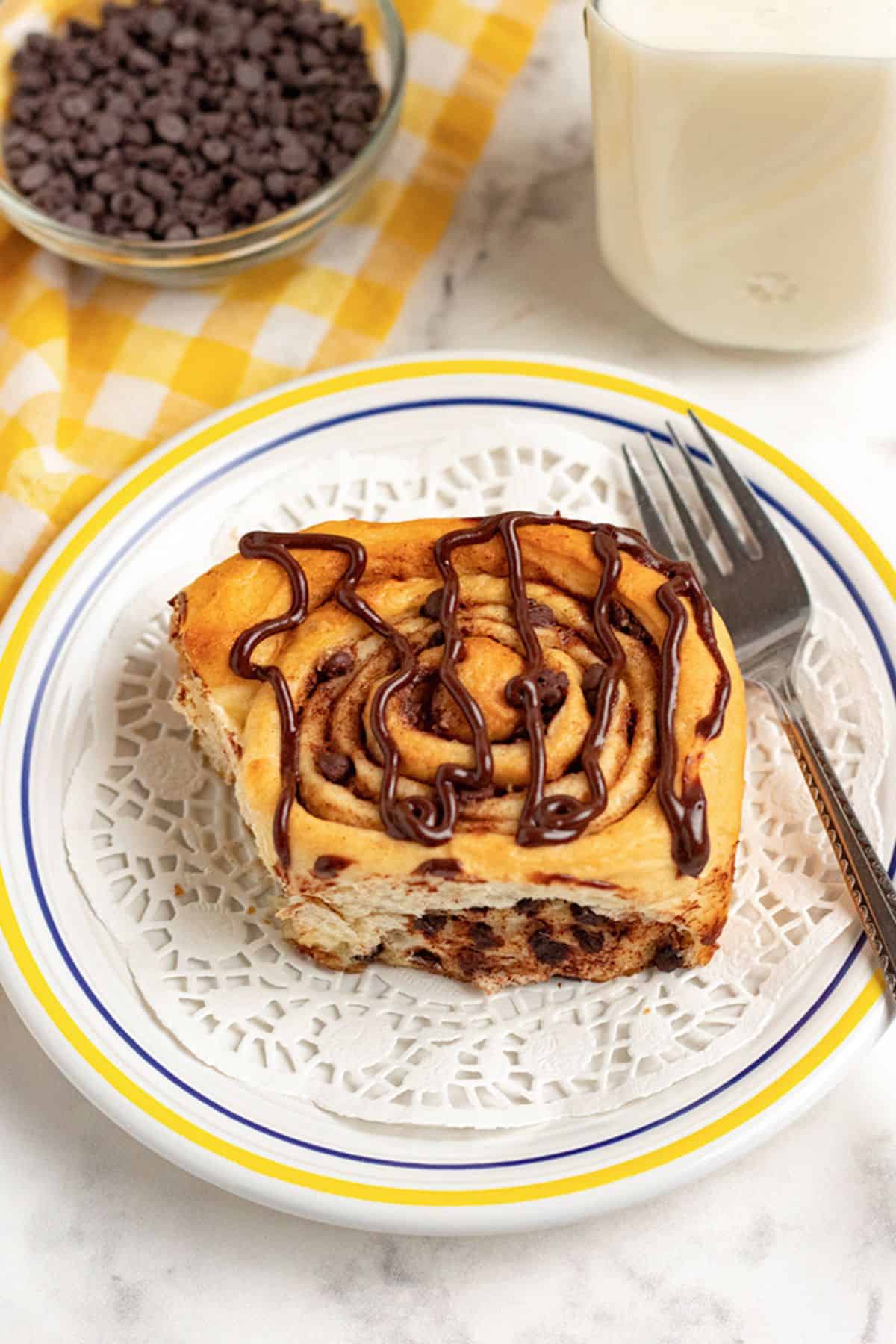 A chocolate chip cinnamon roll with chocolate drizzle sits on a white plate, next to a fork, a glass of milk, and a bowl of chocolate chips atop a yellow checkered cloth.