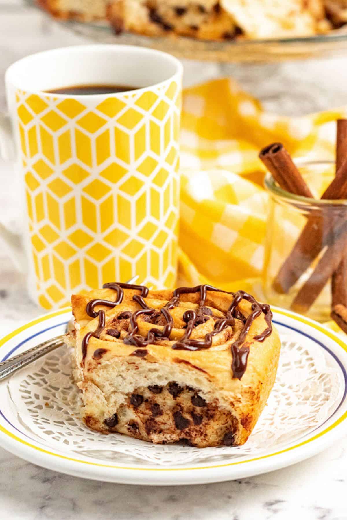 A chocolate chip cinnamon roll drizzled with chocolate sits on a plate next to a cup of coffee, a jar of cinnamon sticks, and a yellow-patterned napkin.
