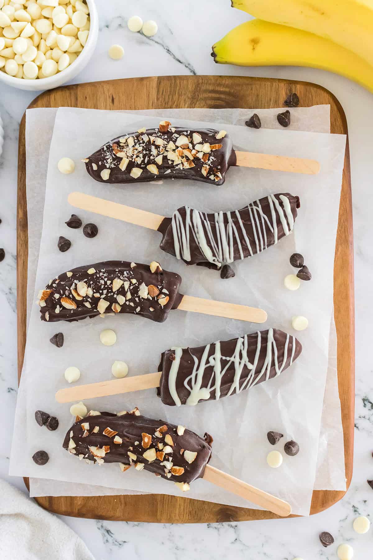 Five frozen chocolate bananas on sticks are arranged on parchment paper atop a wooden board, topped with chopped nuts and white chocolate drizzle. White and dark chocolate chips are scattered around these frozen banana treats.