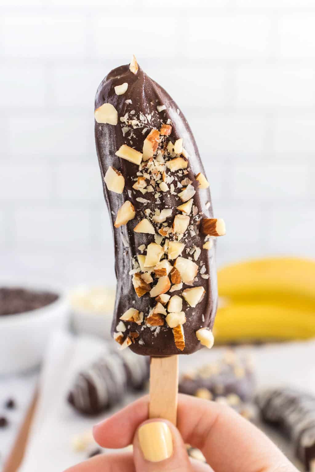 A hand holding a Frozen Chocolate Covered Banana half on a wooden stick, dipped in chocolate and topped with chopped nuts.