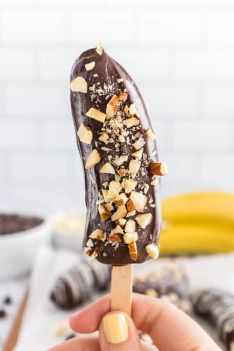 A hand holding a Frozen Chocolate Covered Banana half on a wooden stick, dipped in chocolate and topped with chopped nuts.