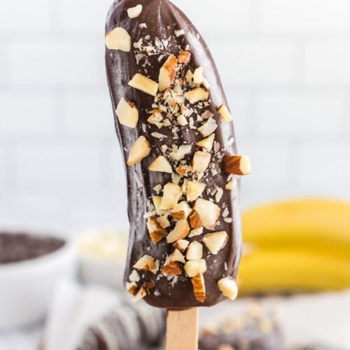 A hand holding a Frozen Chocolate Covered Banana half on a wooden stick, dipped in chocolate and topped with chopped nuts.