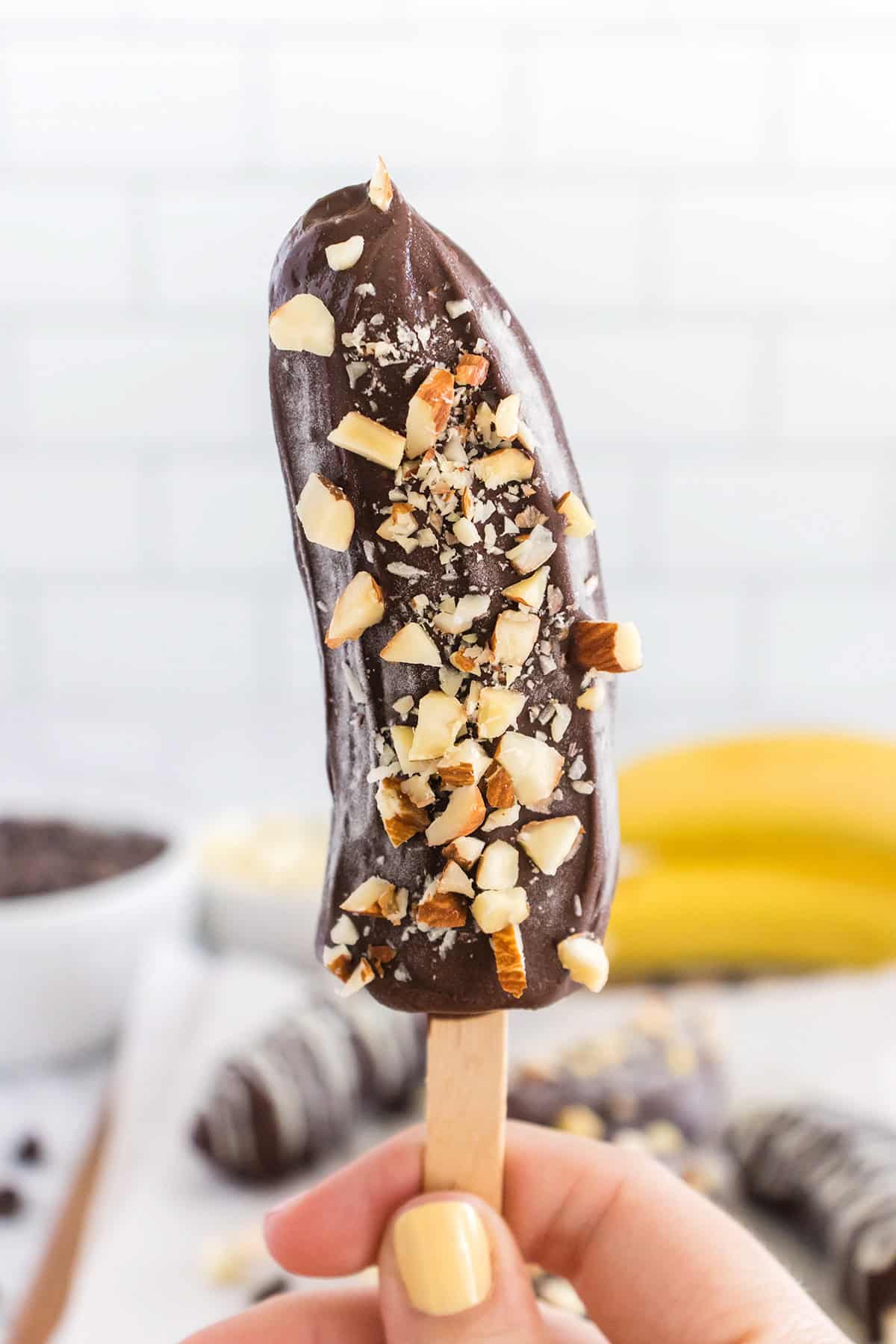 A hand holding a Frozen Chocolate Covered Banana half on a wooden stick, dipped in chocolate and topped with chopped nuts.