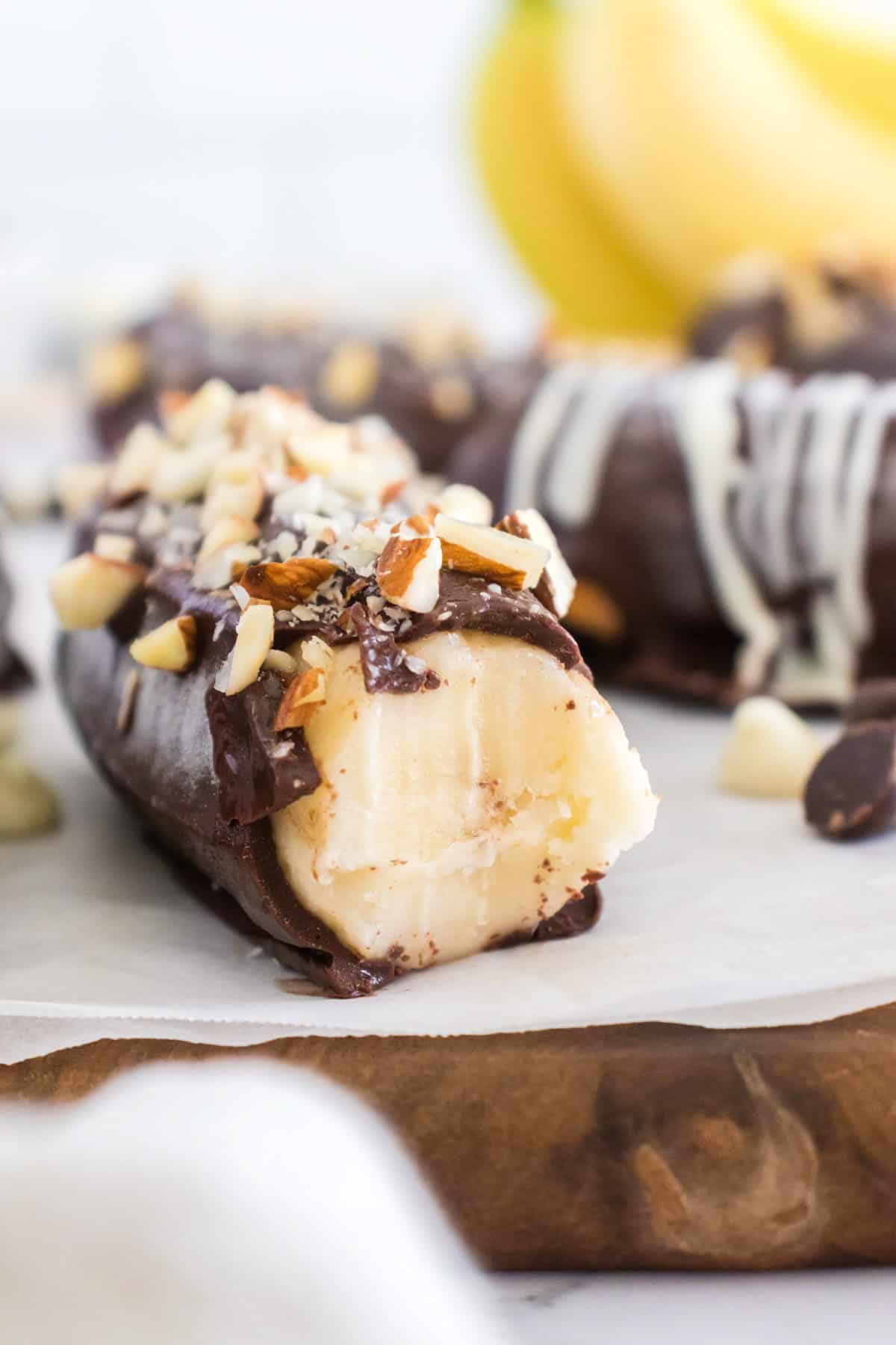 A Frozen Chocolate Covered Banana half is topped with chopped nuts and has a bite taken out, with fresh bananas visible in the background.