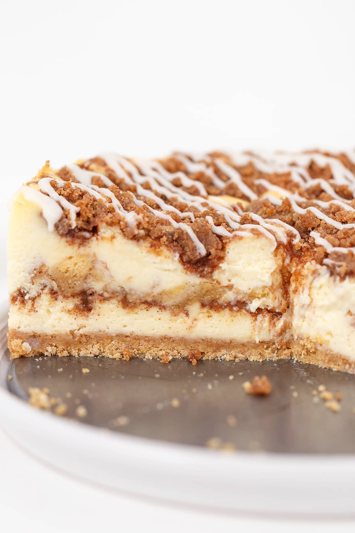 A sliced Honey Bun Cheesecake with a graham cracker crust, cinnamon swirl layers, crumb topping, and white icing drizzle, displayed on a round white plate.