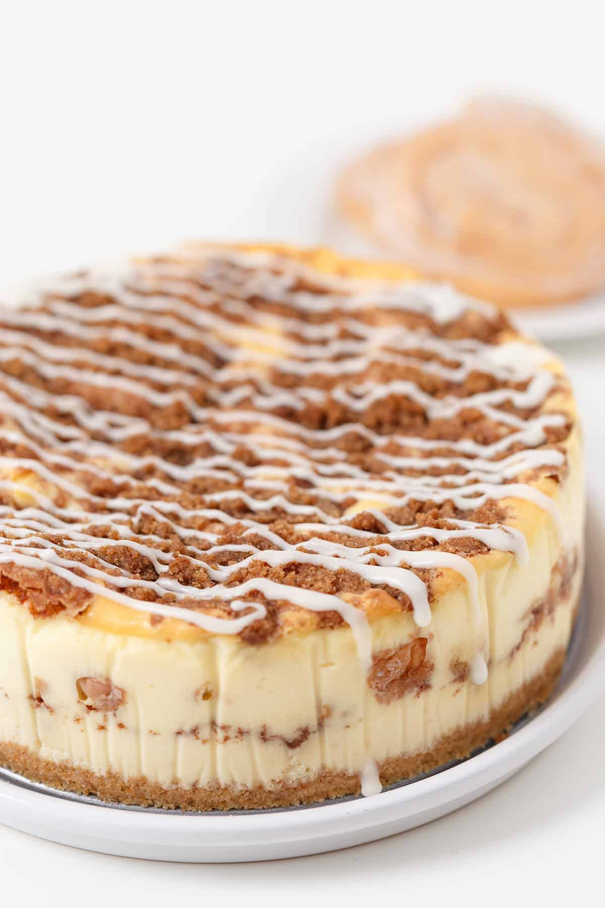 A round Honey Bun Cheesecake topped with cinnamon crumble and white icing drizzle sits on a white plate; a cinnamon roll is blurred in the background.