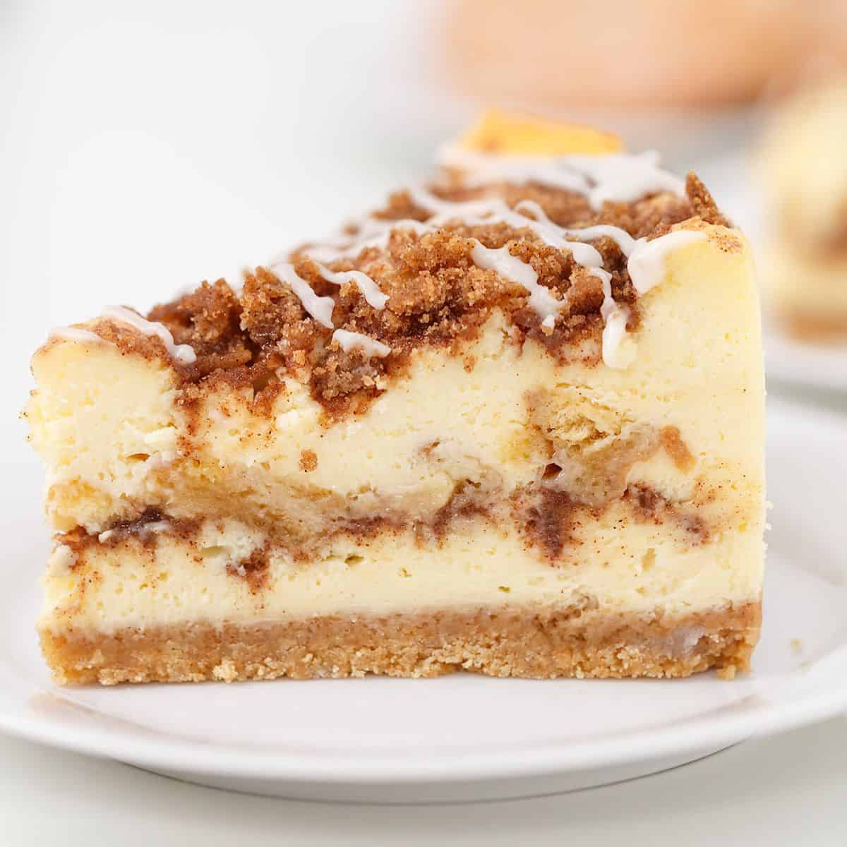 A slice of Honey Bun Cheesecake with a crumbly streusel topping and a drizzle of white icing, served on a white plate.