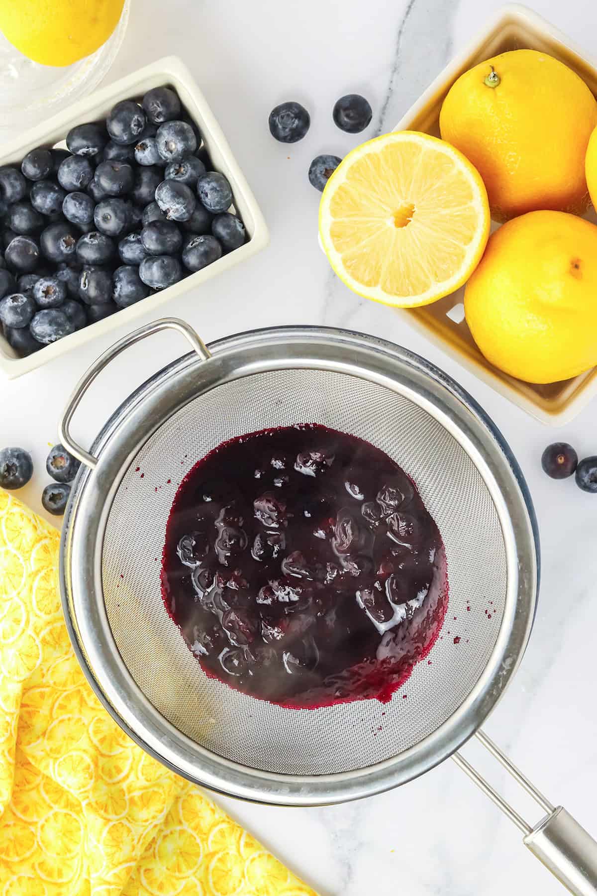 A metal strainer with cooked blueberries sits over a bowl, perfect for a Blueberry Lemon Cake. Fresh blueberries, whole lemons, and a halved lemon are arranged nearby on a white surface.