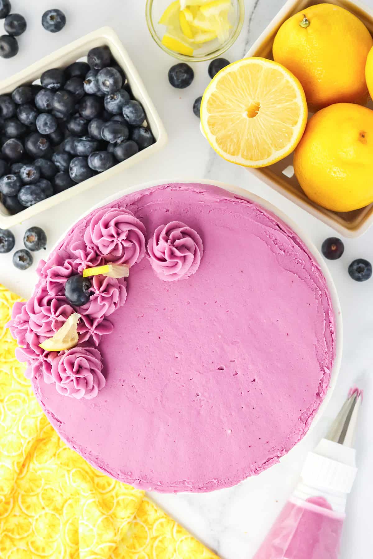 A round Blueberry Lemon Cake with pink frosting, decorated with piped flowers and blueberries, sits next to bowls of fresh blueberries, whole lemons, and lemon slices on a white surface.