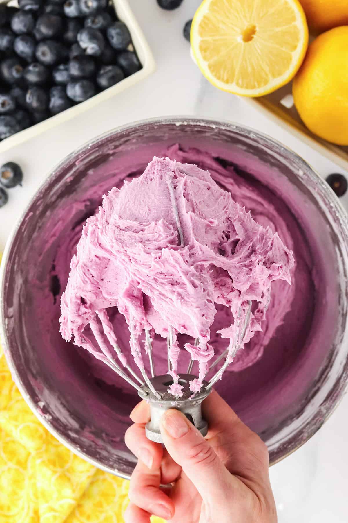 A hand holds a whisk covered in thick, purple frosting above a mixing bowl, with fresh blueberries and halved lemons in the background&mdash;perfect for a homemade Blueberry Lemon Cake.