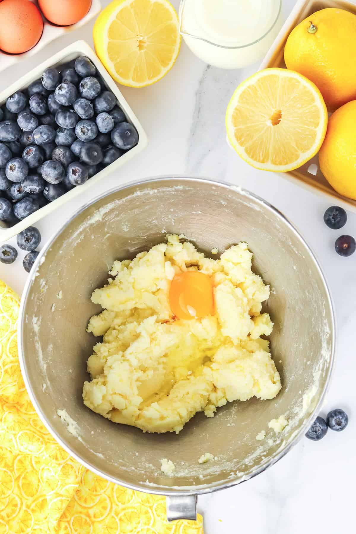 A mixing bowl with butter, sugar, and an egg, surrounded by fresh blueberries, lemons, eggs, and a pitcher of milk&mdash;everything you need to create a delicious Blueberry Lemon Cake on a marble countertop.