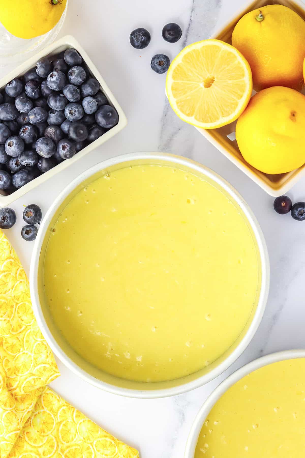 A bowl of yellow lemon batter for Blueberry Lemon Cake is surrounded by fresh blueberries, whole lemons, a halved lemon, and a yellow patterned napkin on a white surface.