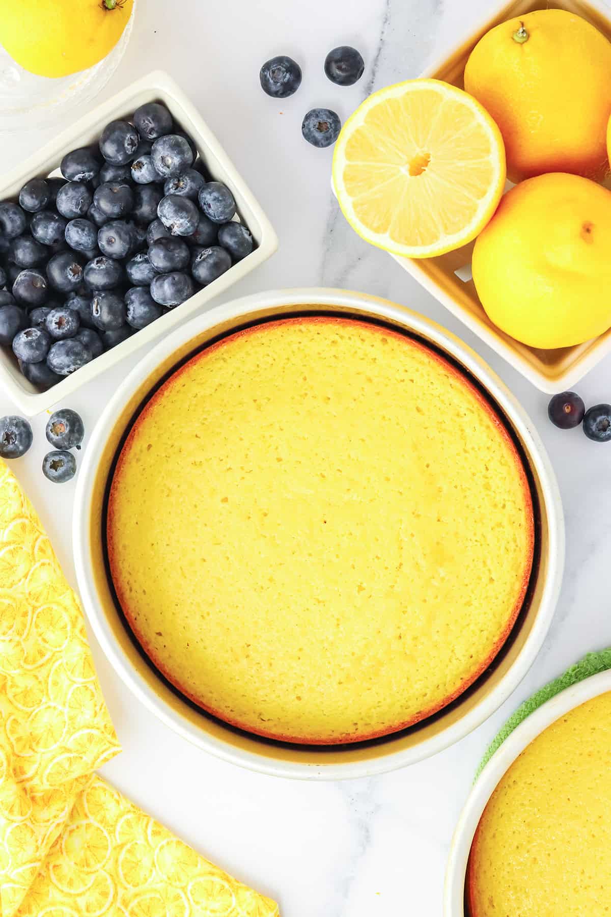 A round, baked Blueberry Lemon Cake in a dish, surrounded by fresh blueberries, whole lemons, a lemon half, and yellow-patterned napkins on a marble surface.