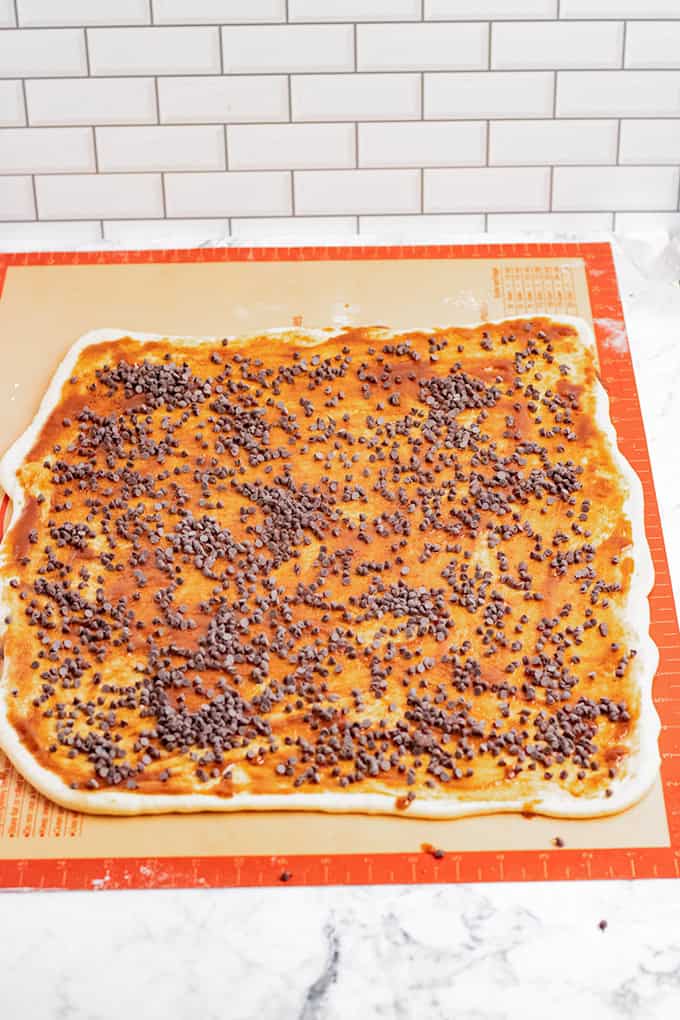 A large rectangular sheet of dough spread with a brown filling and sprinkled with chocolate chips on a silicone baking mat, ready to be rolled into delicious chocolate chip cinnamon rolls.