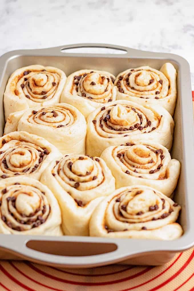 A baking pan filled with unbaked chocolate chip cinnamon rolls, their swirls brimming with rich chocolate chips, sits on a red-striped surface.