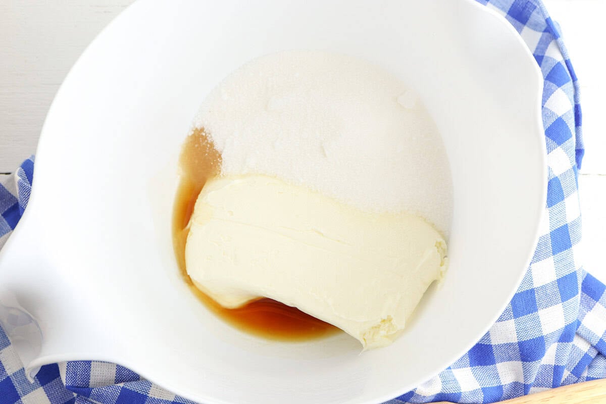 A white mixing bowl containing cream cheese, granulated sugar, and vanilla extract for a delicious Cheesecake Pie sits on a blue and white checkered cloth.