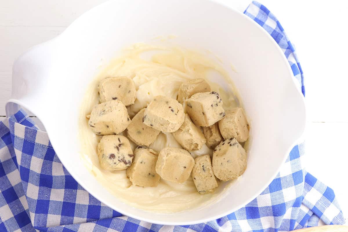 A white bowl on a blue checkered cloth contains raw cookie dough cubes and cake batter, perfect for making a decadent cookie dough cheesecake or a classic cookie dough pie.