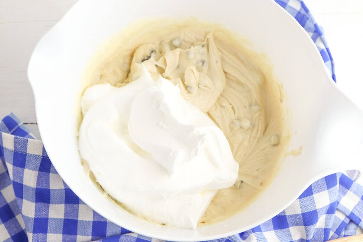 A white mixing bowl with Cookie Dough Cheesecake filling and a large dollop of whipped cream on top, set on a blue and white checkered cloth.