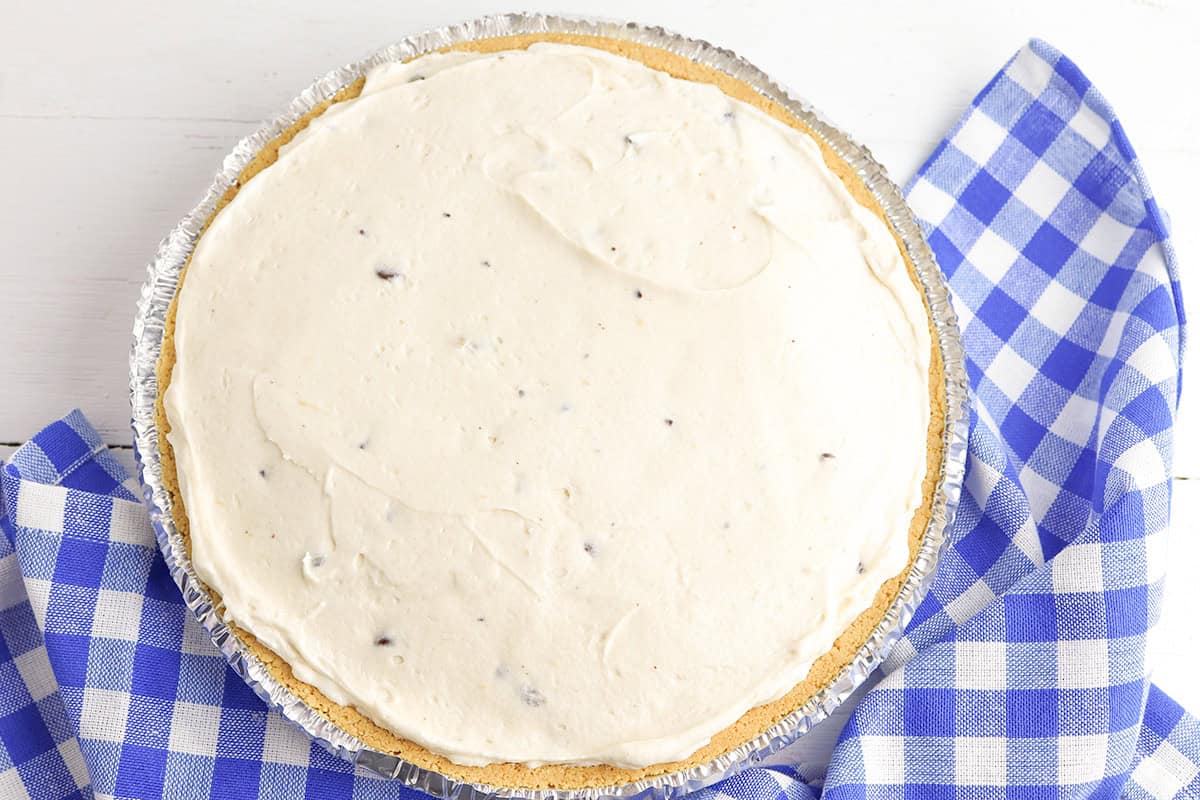 A creamy cheesecake pie with a graham cracker crust sits on a blue and white checkered cloth.