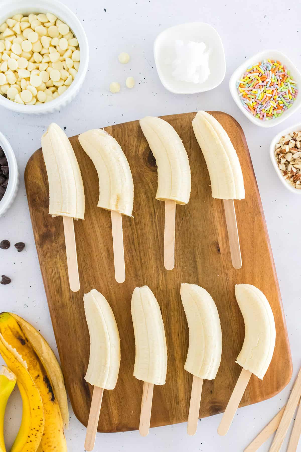 Eight halved bananas on wooden sticks are arranged on a cutting board, ready to be transformed into Frozen Chocolate Covered Bananas with bowls of white chocolate chips, sprinkles, chopped nuts, and coconut oil.